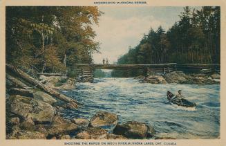 A man in a canoe navigating a river full of rapids. Two people look on from a log bridge.