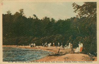 Many people gather by a water's edge with a forested area in the background.