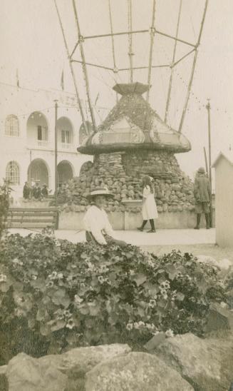 Picture of woman standing near a garden with large rock monument and building in background. 
