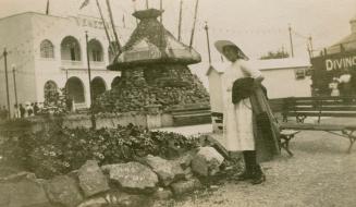 Picture of woman standing near a garden with large rock monument and building in background. 