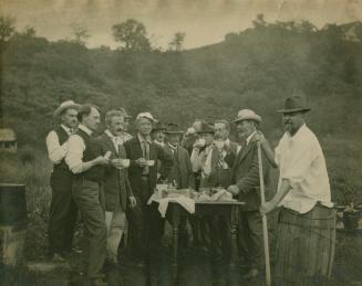 A photograph of twelve men standing around a table in the middle of a farm. They are all middle ...