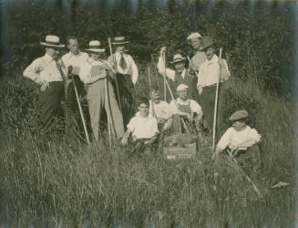 A photograph of eleven people standing and sitting in a grassy field. They are wearing hats, dr ...
