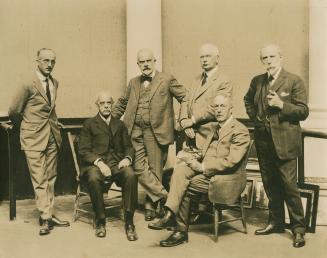 A photograph of six men sitting and standing in a room with framed pictures propped against the ...
