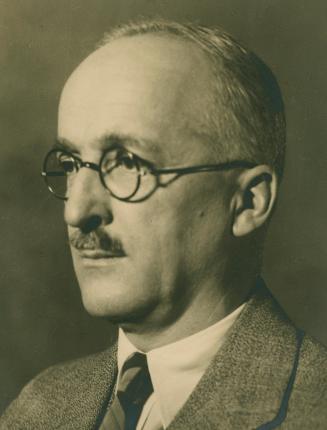 A photograph of a middle-aged man from the shoulders up. He is wearing a suit and tie, glasses  ...