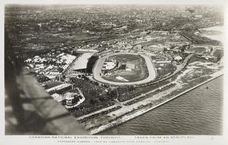 Aerial shot of a grand stand, track and a roller coaster in an amusement park by a lake. B & W.