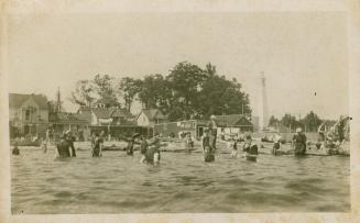 Black and white picture of people wading in water beside a beach with houses.