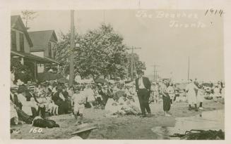 Black and white picture of a crowd of people on a sandy beach with houses to the left.