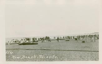 Black and white picture of a crowd of people on a sandy beach to the right of a large body of w ...