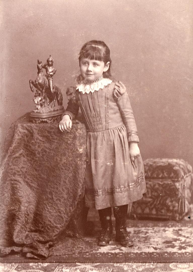 A posed photograph of a young girl standing beside a statue of a knight on a horse. The statue …