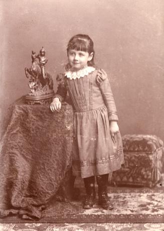 A posed photograph of a young girl standing beside a statue of a knight on a horse. The statue  ...