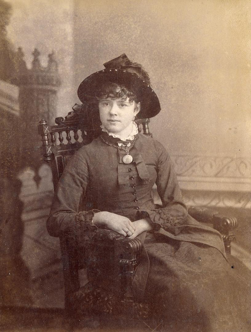 A posed cabinet card photograph of a young woman sitting in an armchair. She is wearing formal …