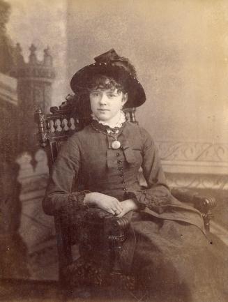 A posed cabinet card photograph of a young woman sitting in an armchair. She is wearing formal  ...