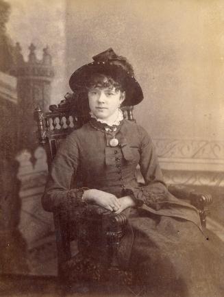A posed cabinet card photograph of a young woman sitting in an armchair. She is wearing formal …