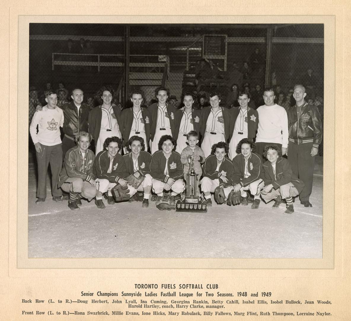 A photograph of a women's softball team posing in front of a trophy. They are sitting and stand…