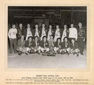 A photograph of a women's softball team posing in front of a trophy. They are sitting and stand ...