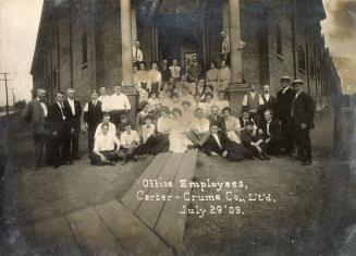 A photograph of approximately fifty men and women sitting and standing in front of a large bric ...
