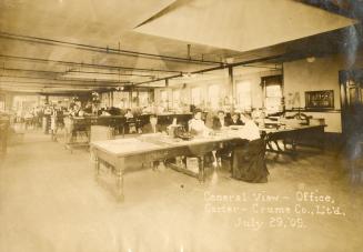 A photograph of the interior of an office, with several men and women sitting at long tables an ...