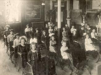A photograph of approximately twenty young students, boys and girls, sitting at wooden desks in ...