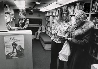 Interior view of a bookmobile with woman looking at a book with a librarian. 