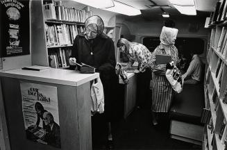 Shows a number of people using a Bookmobile. An older woman in a rain hat looks at a hardcover  ...