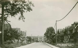 Black and white photograph of school at the end of a street lined with two story houses.