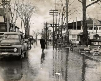 Picture of a woman walking down flooded street with stores on both sides and a truck parked on  ...