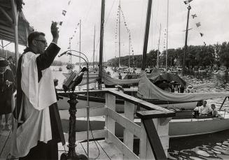 Picture of minister standing on dock blessing several boats in a lagoon. 