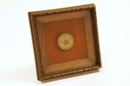 A round metal disk with writing and a coat of arms on it, mounted in a wooden frame.