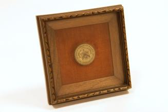 A round metal disk with writing and a coat of arms on it, mounted in a wooden frame.