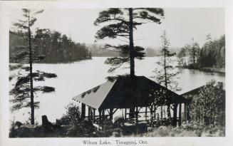 Black and white photograph of covered structure in front of a lake in the wilderness.