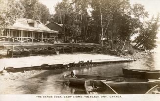 Black and white photograph of boats at a shoreline in front of a large cottage.