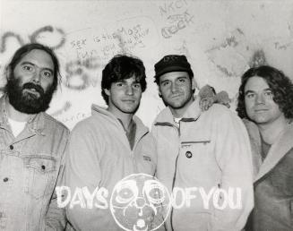 A photograph of a musical group posing in front of a wall with graffiti on it, and the band nam ...