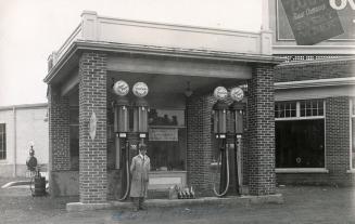 Black and white photograph of a man standing in front of gasoline pumps in a service station.