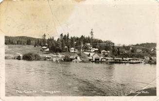 Black and white photograph of cabins beside a lake in a wild, wooded area.
