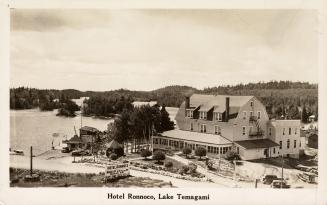 Black and white photograph of a hotel in a wild, wooded area beside a lake.