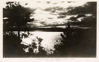 Black and white photograph of the sun going down on a lake in the wilderness. 