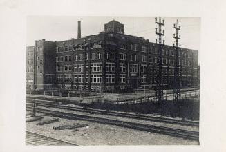 Black and white photograph of a four story hospital building with railway tracks in the foregro ...