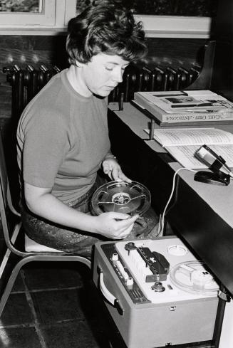 A woman sitting at a desk winds a large cassette tape. 