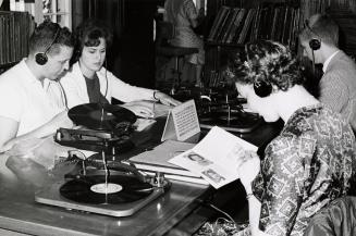 Four people sit at a table in a music library listening to records. 