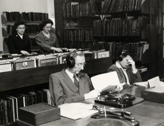 Photo of two people in a library listening to records at a table and two others standing lookin ...