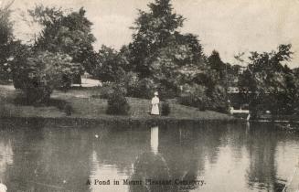 Black and white postcard of a pond surrounded by trees. The caption on the bottom front states, ...