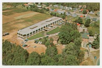 Color, aerial photograph of a large hospital building beside a small town in the wilderness.