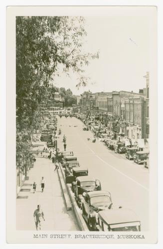 Black and white photograph of the main street in a small town. Cars are parked bumper to bumper ...