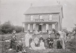 A photograph of approximately twelve people posing on a lawn in front of a two-story house. The ...
