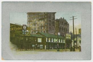 Picture of a street with buildings on one side and large building in background. Silver border …