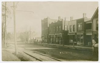 Black and white photograph of small town street with stores on the east side of it.
