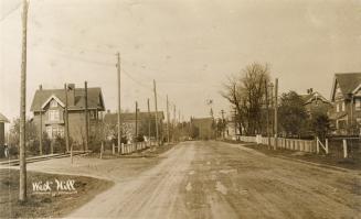 Picture of wide dirt street with houses on both sides and church in distance. 
