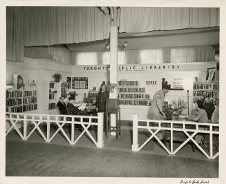 Photo of library booth with men reading at tables and talking to librarian at a desk.