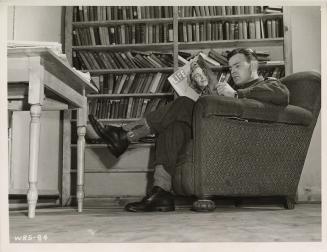 Photo of a man seated in a chair reading a magazine in a library.