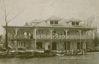 Black and white photograph of many boats and canoes docked at a two story club house.